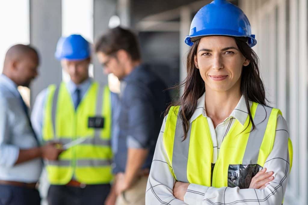 woman in hi-vis vest and blue hard hat standing with her arms crossed, smiling, with three men talking in background