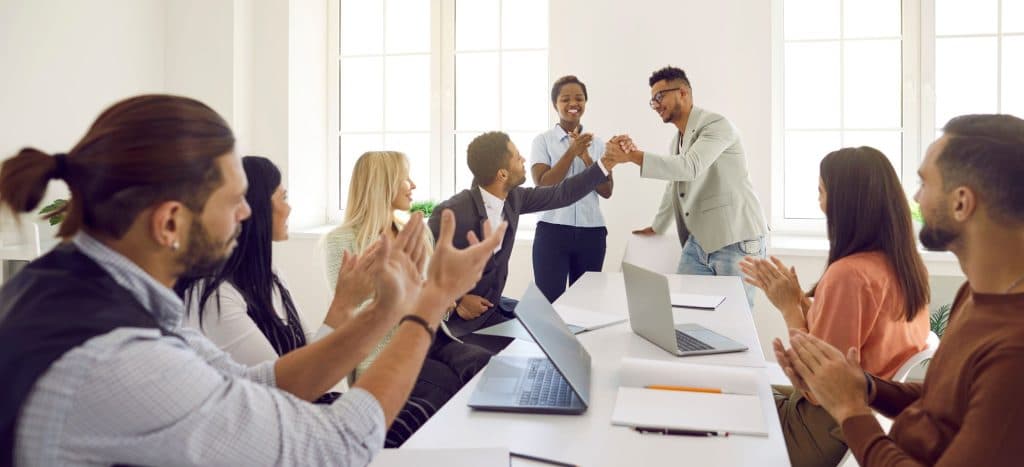 Friendly and diverse employees congratulate man on business achievements, excellent work results or promotion. Business people applaud and cheers during business meeting in bright modern office