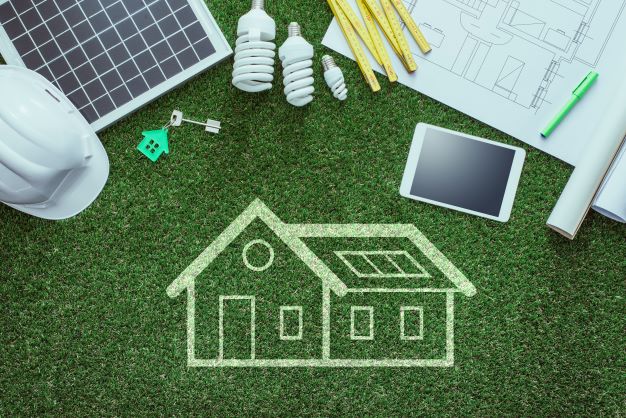 white outline of house on green turf next to hard hat, solar panel, lightbulbs, key, blueprints, ruler, table, and pen