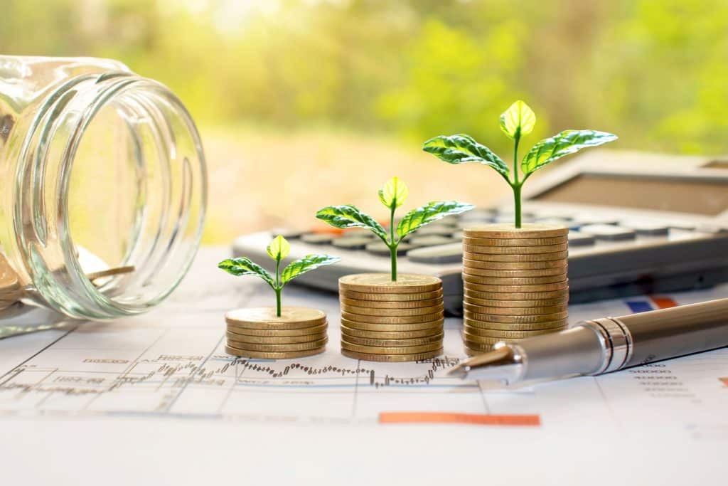 little plants sprouting out of three small towers of gold coins next to a jar on its side, a calculator, and a pen